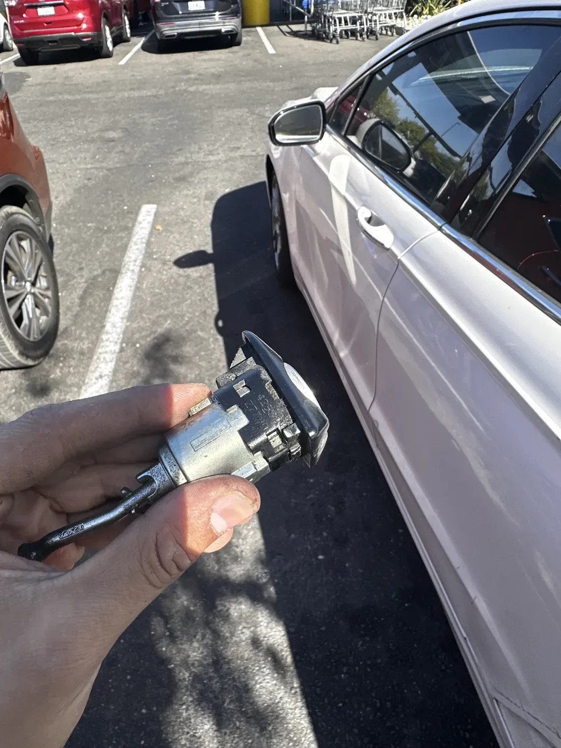 A removed car door lock cylinder held in a hand next to a white vehicle in a parking lot