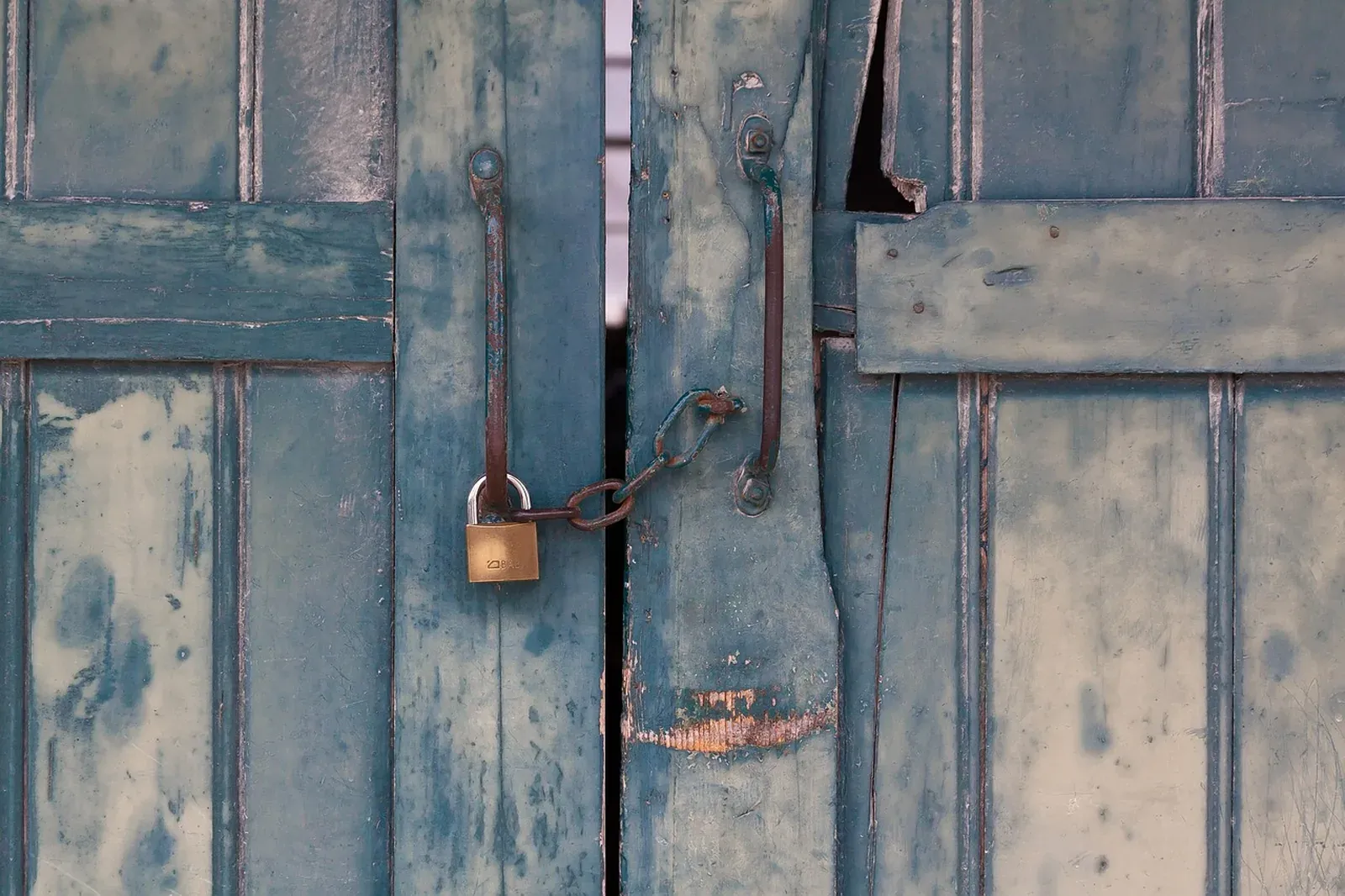 Brass padlock on a short chain and iron handles across weathered teal-painted barn doors